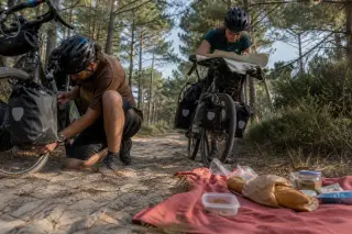 Two cyclists take a break on a sandy forest trail in Gironde, one checking a bike tire, the other reading a map, with picnic items nearby and sunlit pine trees all around.