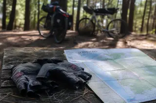 Worn cycling gloves and paper map on a wooden bench in a pine forest, with bikes resting against trees in the blurred background, suggesting a peaceful route planning break.
