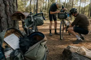 Two cyclists at the edge of a pine forest in the Landes, one checking a GPS, another adjusting panniers beside slightly dusty gravel and VTC bikes leaned against a wooden fence in soft afternoon light.