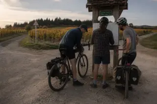 A family takes a quiet break along a wooded bike trail, organizing gear and filling water bottles near two touring bikes and a signpost.