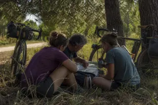Three cyclists consult a map during a break under pine trees beside a remote Gironde cycling trail, their bikes resting nearby in gentle afternoon light.