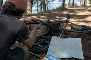 Casual cyclist repairing a flat tire in pine forest near Arcachon, local map unfolded on ground, soft afternoon light.