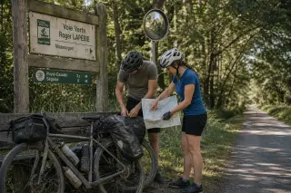 Cyclotourists consult a map at a rustic trail sign along a wooded voie verte, bikes resting nearby, in soft late afternoon light.
