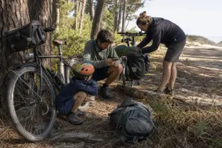 Family with used bicycles pauses beside a pine forest bike trail in the Gironde, child watching parent check smartphone, afternoon light filtering through trees.