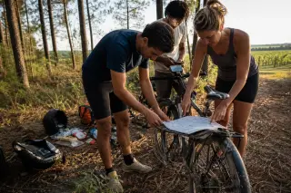 Cyclists consulting a map at the edge of a pine forest, bikes dusty and gear spread out, late afternoon sunlight streaming through the trees.